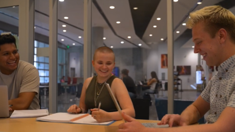 Three students laughing while talking to each other at a table with laptops and notebooks in front of them with windows and the Canyon Cafe in ENR2 in the background. 